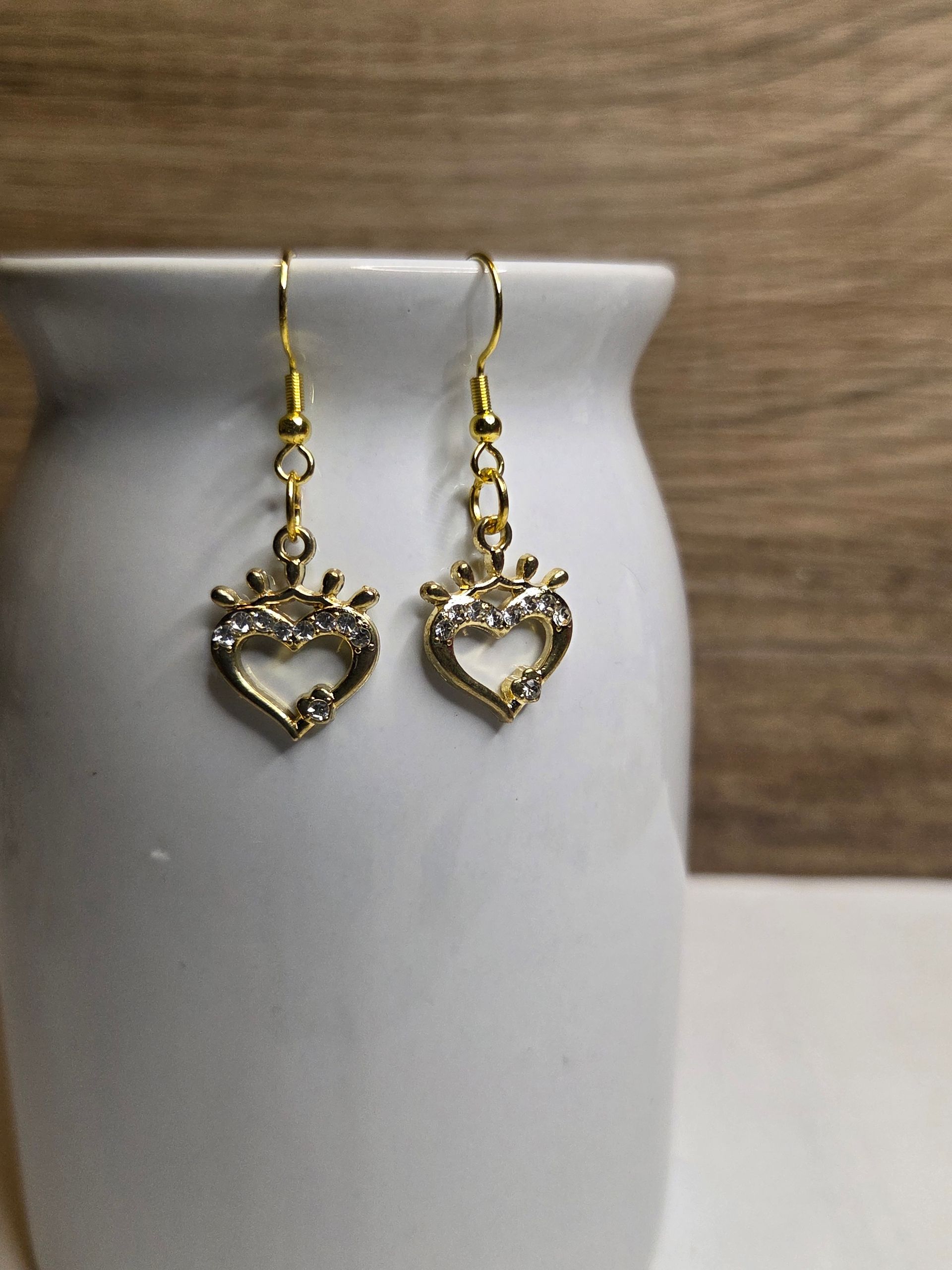 Aan een witte vaas hangen goudkleurige oorbellen, elk met een hartvormig ontwerp en een kroontje.