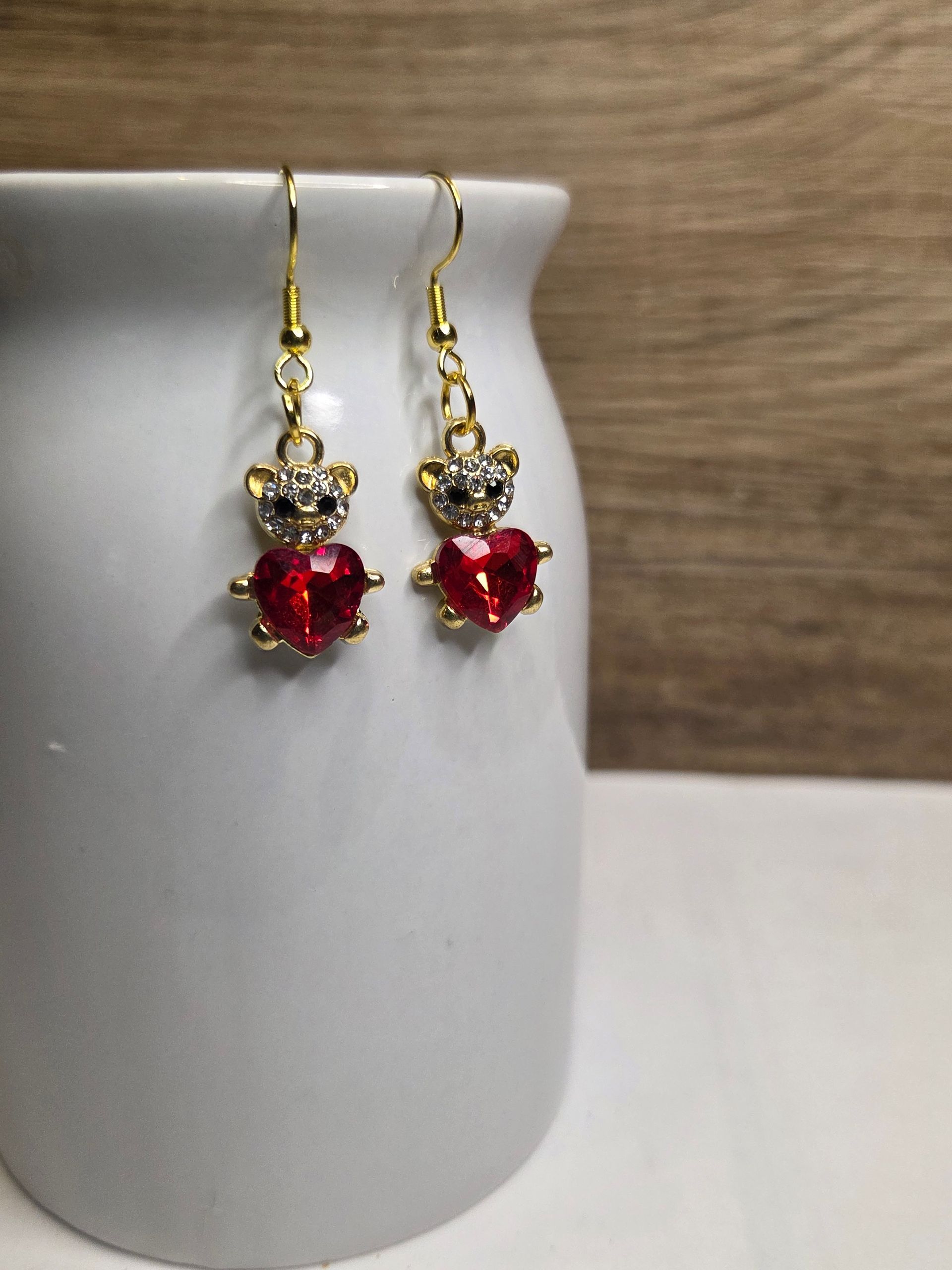 Gouden oorbellen in de vorm van een teddybeer met rode hartvormige edelstenen, hangend aan een witte vaas.