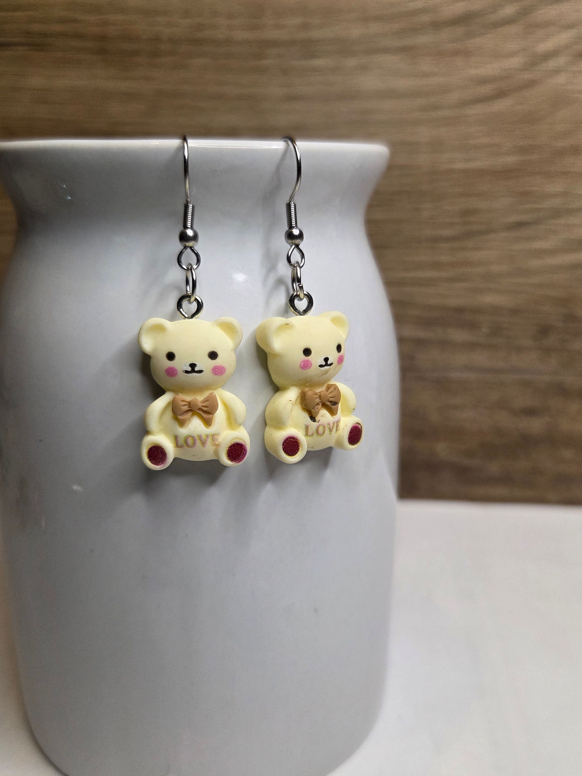 Crèmekleurige oorbellen in de vorm van een teddybeer met rode pootjes en bruine strikjes, hangend aan zilveren haakjes.