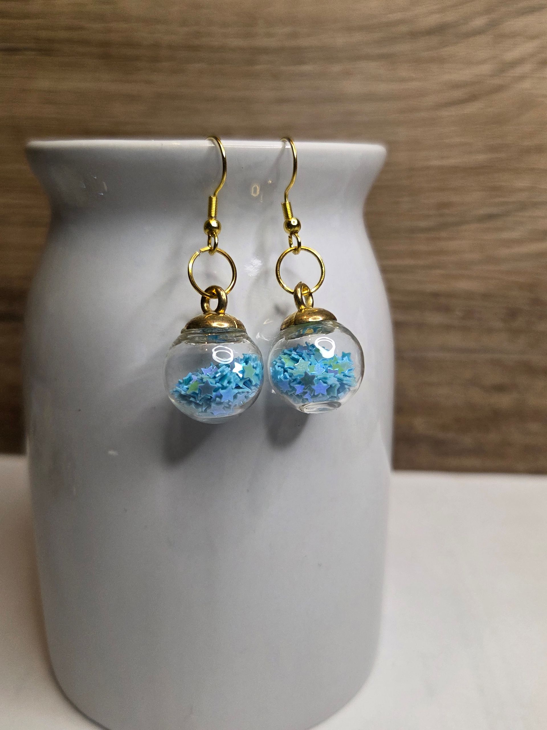 Oorbellen met transparante bolletjes gevuld met blauwe glitter, hangend aan een witte vaas.