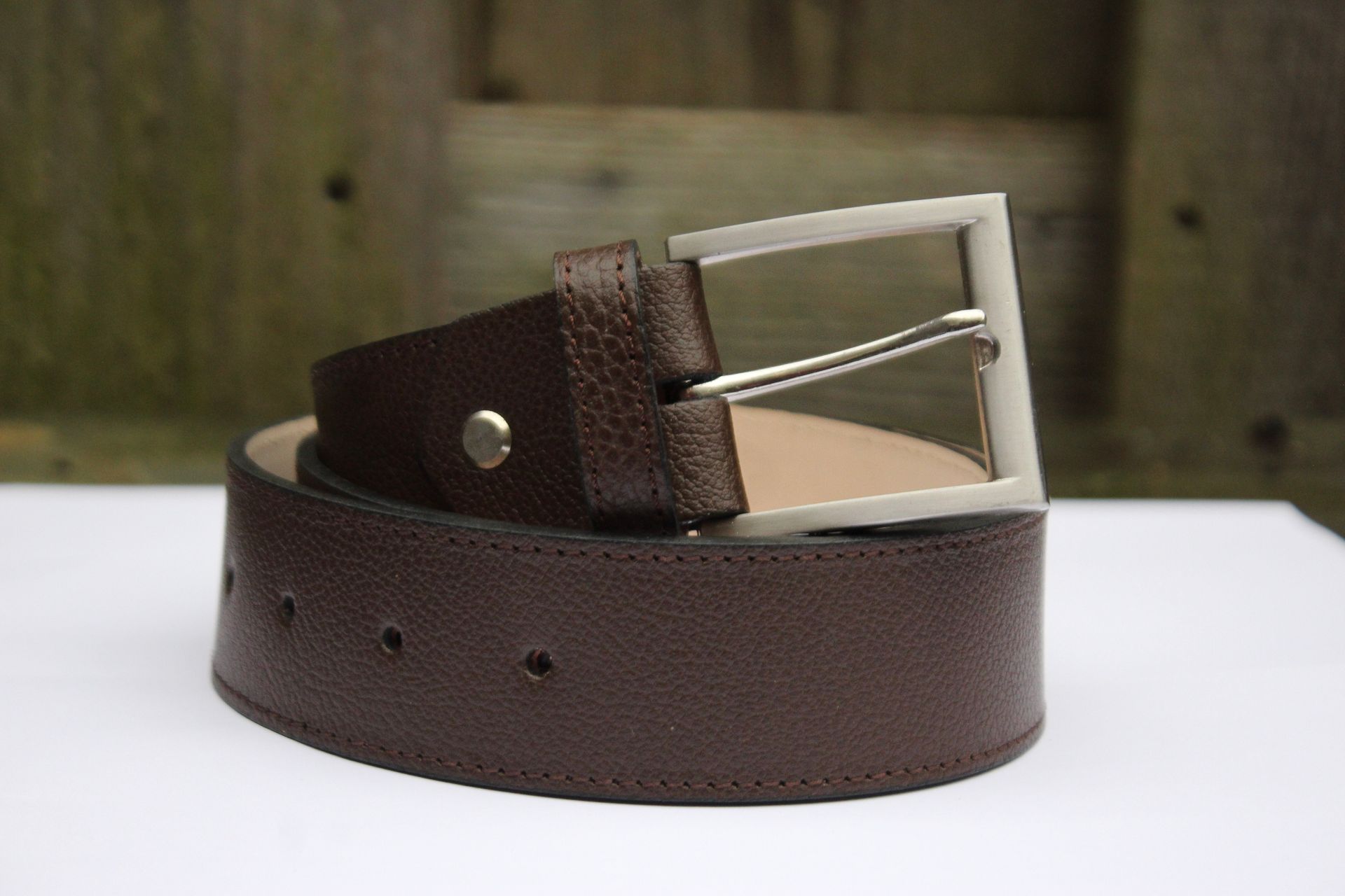 Bruine leren riem met zilveren gesp, opgerold op een witte ondergrond met een houten achtergrond.