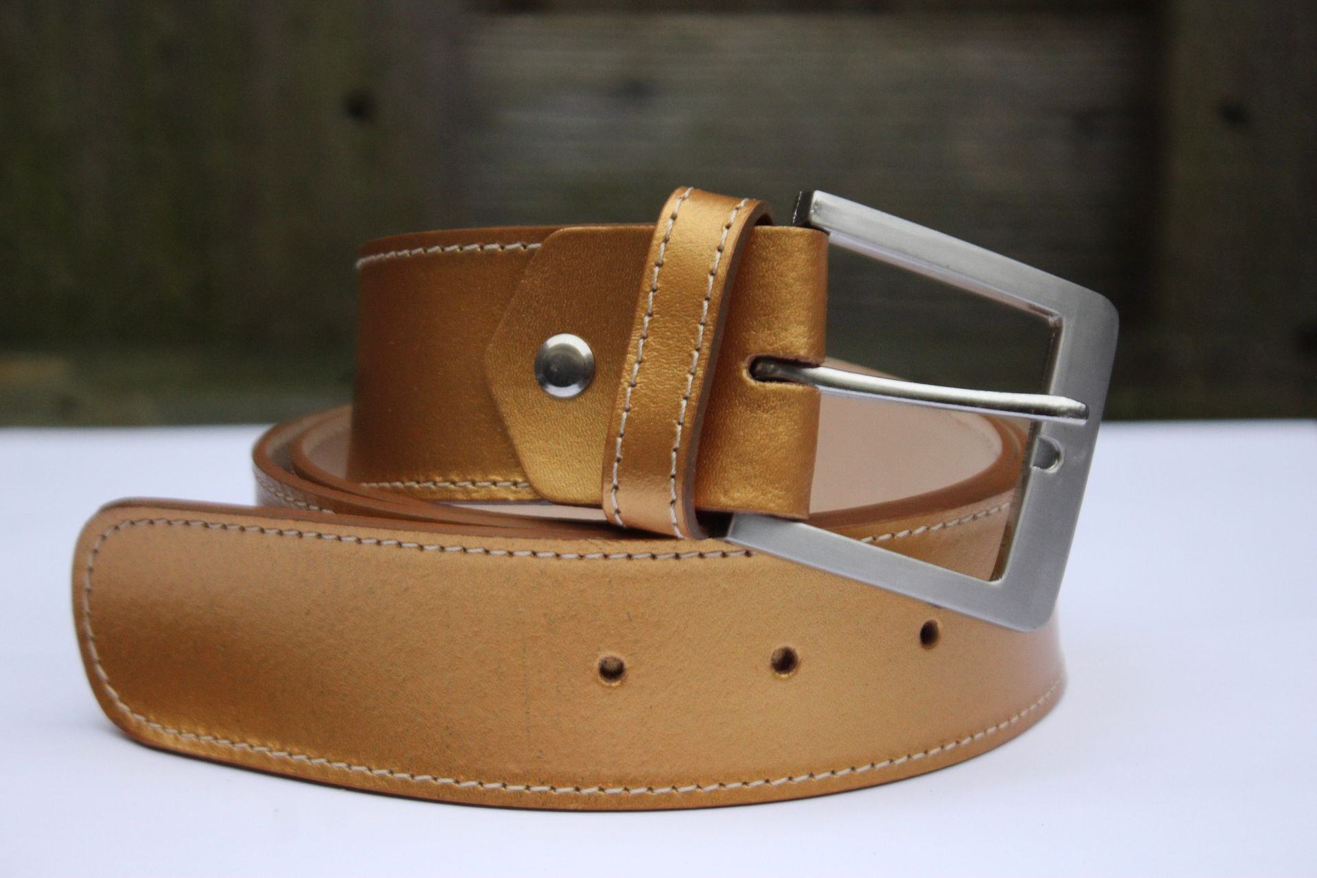 Goudkleurige leren riem met een zilveren vierkante gesp, opgerold op een witte ondergrond met een wazige houten achtergrond.