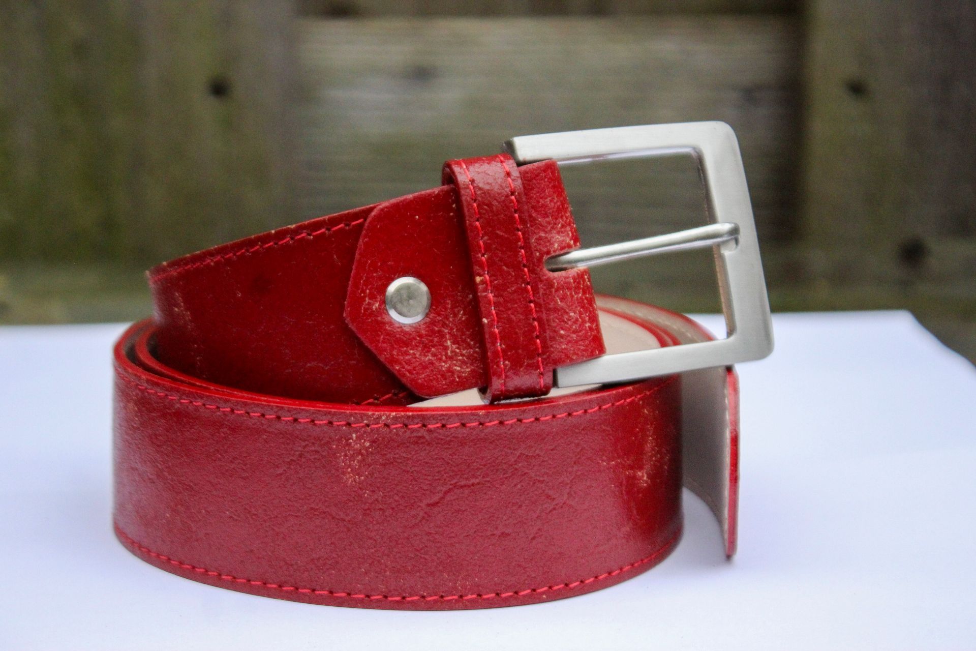 Rode leren riem met zilveren gesp, opgerold op een witte ondergrond met een houten achtergrond.