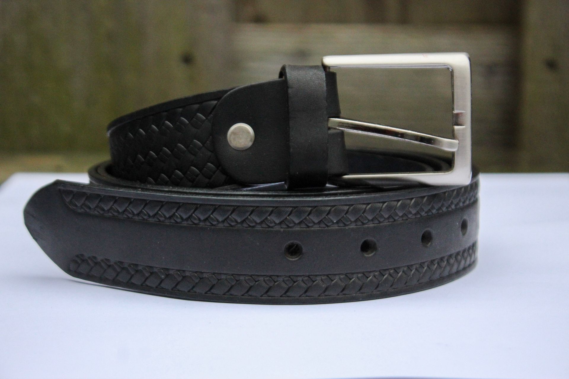 Zwarte leren riem met zilveren gesp, opgerold op een witte ondergrond met een houten achtergrond.