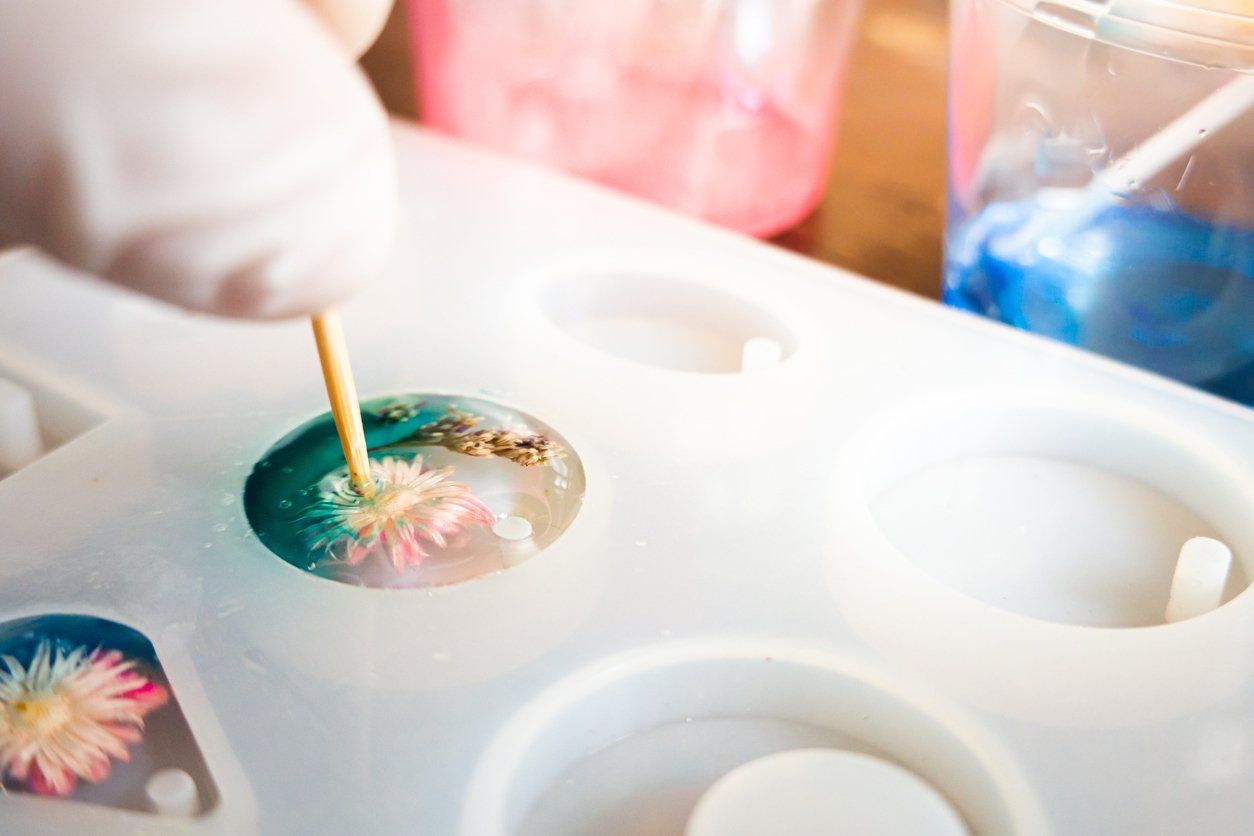 Een persoon gebruikt een tandenstoker om bloemen te schikken in een met hars gevulde mal; kleurrijke hars op de achtergrond.