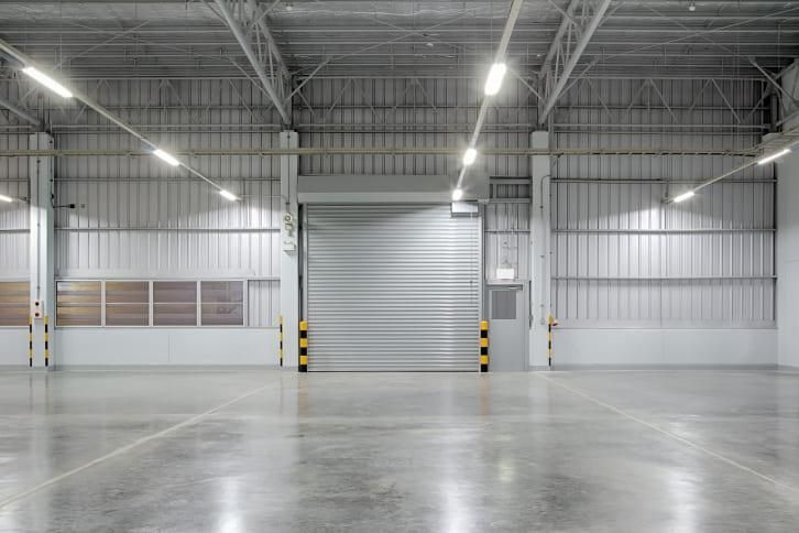 Empty warehouse interior with a closed overhead door. Metal walls, concrete floor, overhead lights.