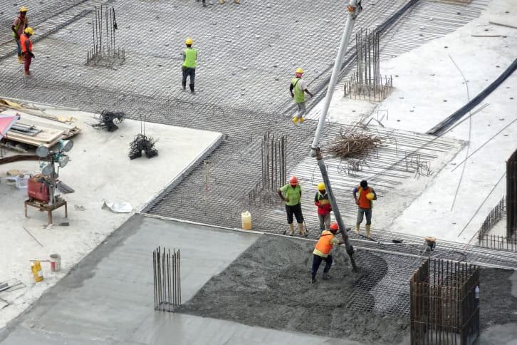 Workers in safety vests pour wet concrete onto a reinforced steel construction site from a boom pump.