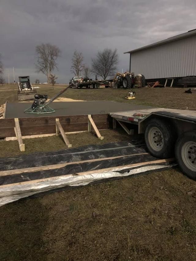 Construction site with a concrete slab, wooden forms, and a trailer. A cloudy sky is overhead.