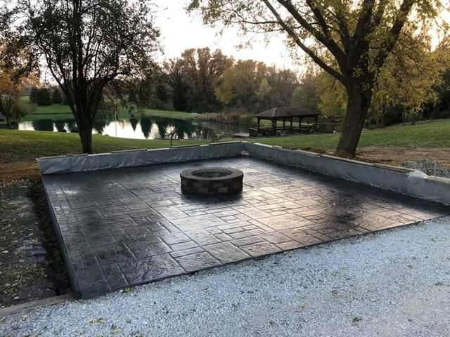 Concrete patio with fire pit, gravel border, and trees, overlooking a pond.