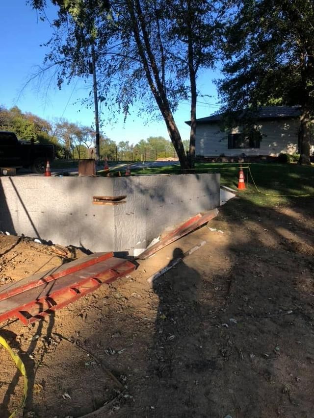 Construction site with concrete wall, dirt, orange cones, and a house in the background.