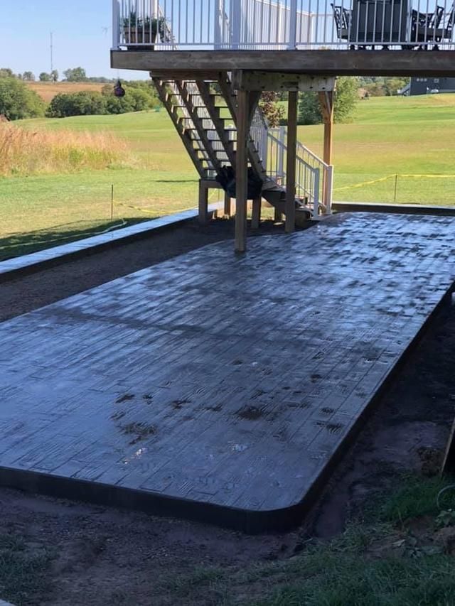 A freshly poured concrete patio under a deck.  A grass lawn and steps leading up from the patio are visible.