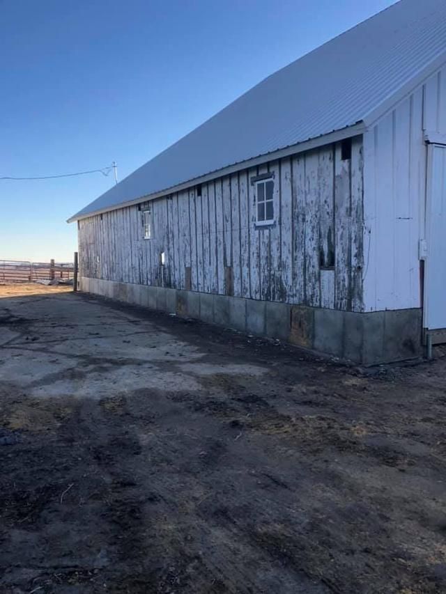 White barn with weathered paint and metal roof on a dirt lot under a clear blue sky.