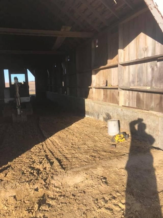 Interior of a barn with dirt floor, beams, and weathered wooden walls. Sunlight casts a shadow.