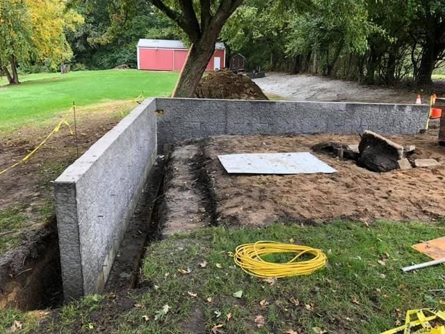Concrete foundation walls under construction in a grassy yard, with a trench, a tree, and a red shed in the background.