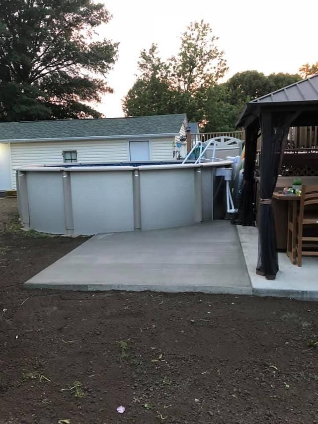 Above-ground pool on concrete pad next to a gazebo and building, set in a yard.