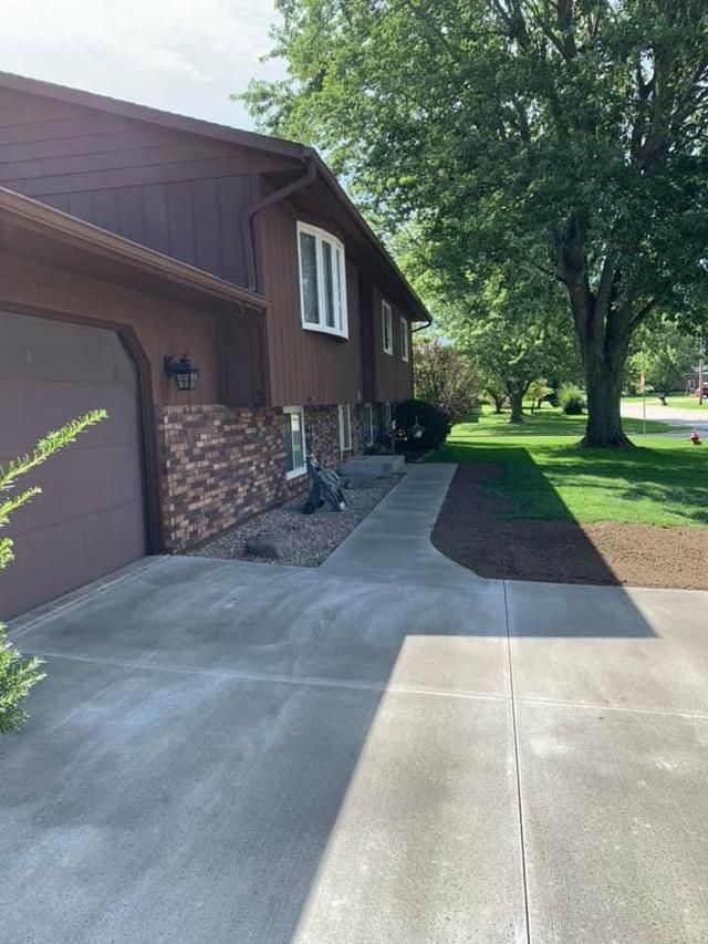 Brown house with brick accents, driveway, and sidewalk leading to the front door. Lawn and trees surround.