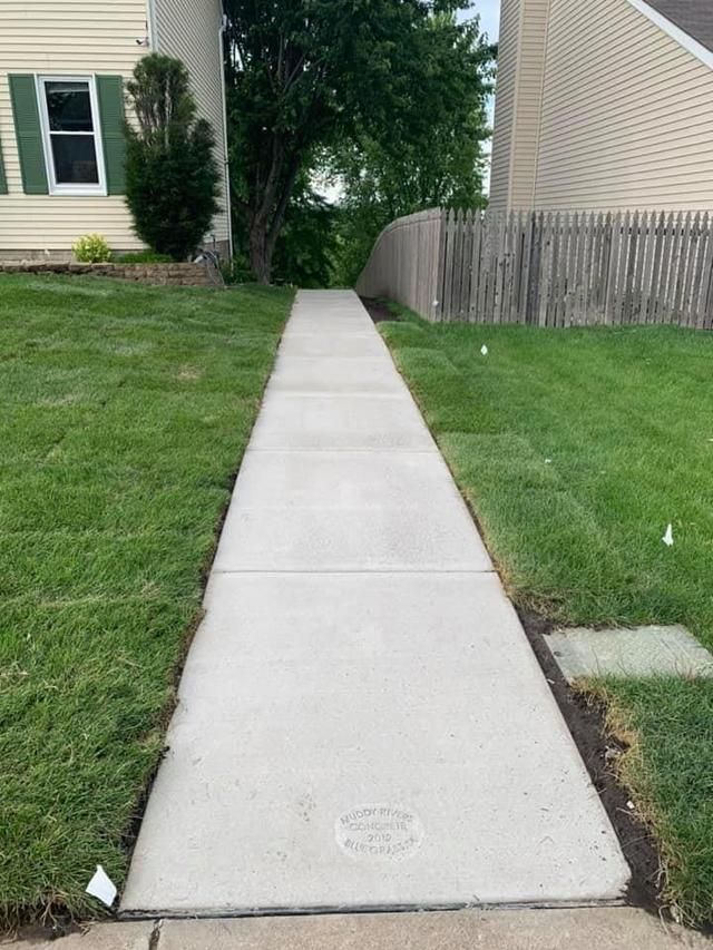 A concrete sidewalk runs between green lawns, leading towards trees and a fence. Houses on either side.