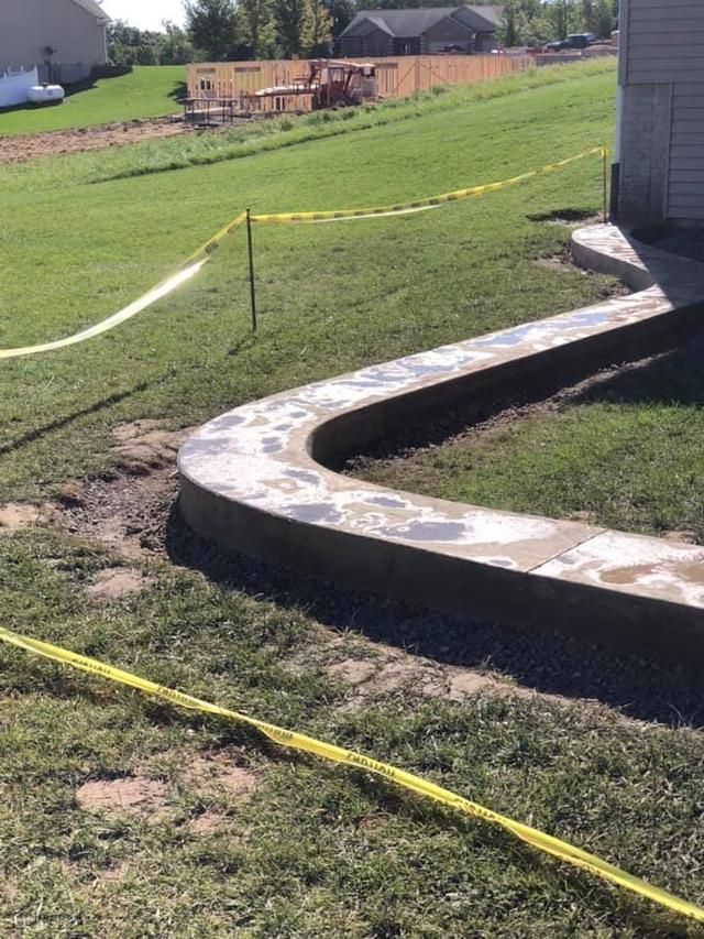 Newly poured concrete pathway curving through a grassy yard, yellow caution tape nearby.