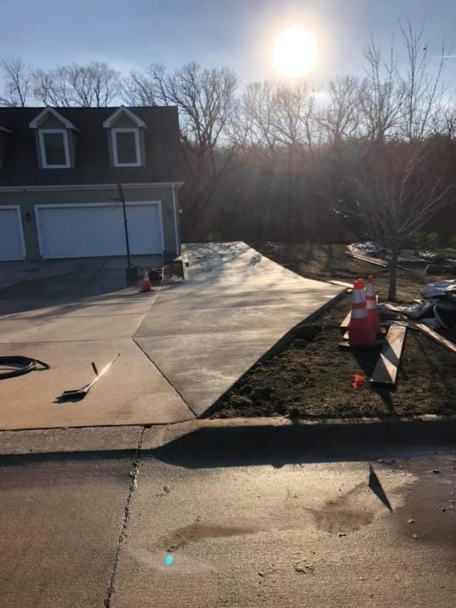 Newly poured concrete driveway in front of a house, orange cones mark boundaries, sun shining brightly.