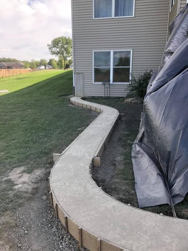 Curved concrete walkway next to a two-story building and grass. Forms visible along the edges.