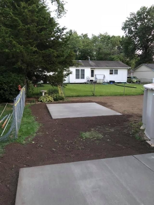 Newly poured concrete pad in a yard, with a house in the background. Freshly tilled earth surrounds the pad.