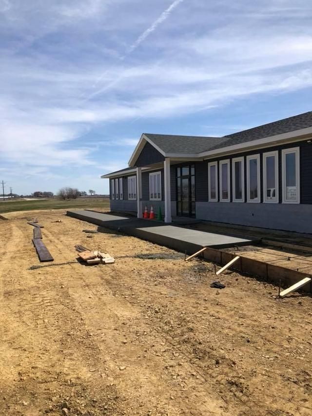 Building exterior under construction; gray siding, concrete patio, dirt, clear sky.