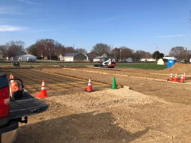 Construction site with an excavator, orange cones, and rebar. Gravel and dirt ground, blue sky.