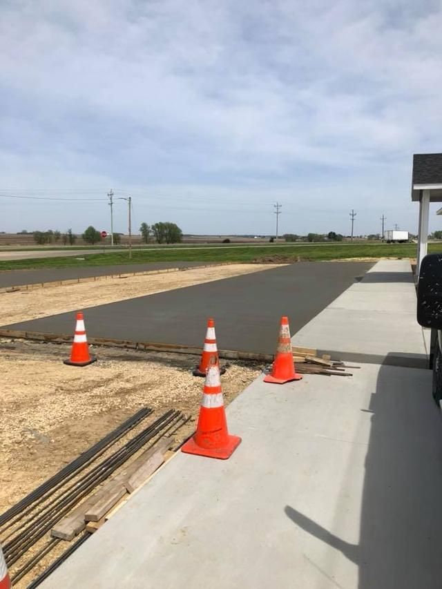 Freshly poured concrete area, bordered by orange cones and a sidewalk, with a rural landscape in the background.