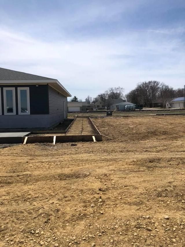 A partially constructed building with a dirt yard, raised garden beds, and distant houses under a blue sky.