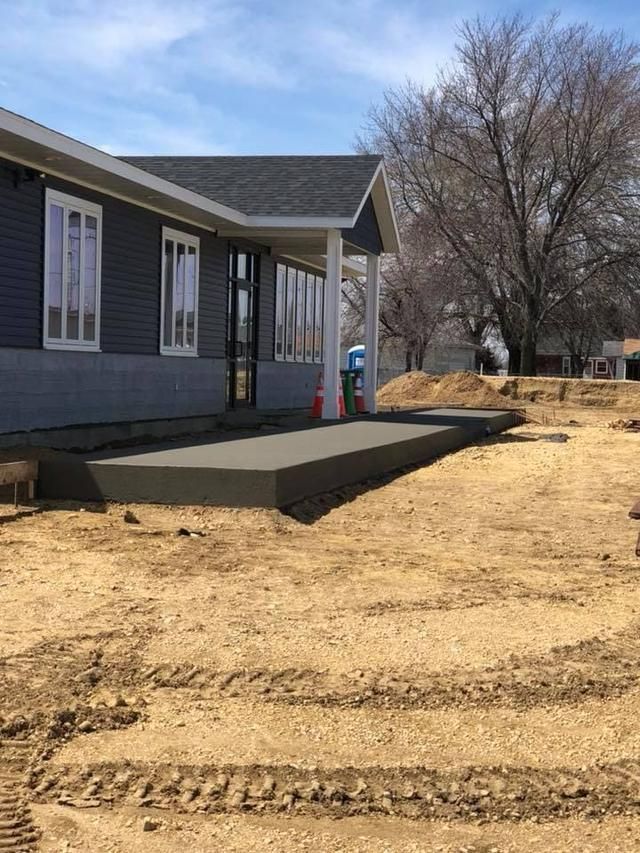 A new concrete porch with white pillars stands outside a dark gray building on a construction site with dirt ground.