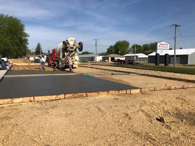 Concrete being poured on a street construction site; a cement truck, workers, and a building are visible.