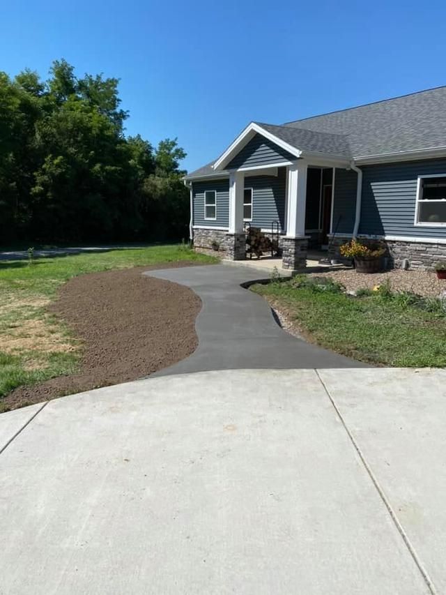 Asphalt walkway leading to a blue-sided house with a porch and gray roof on a sunny day.