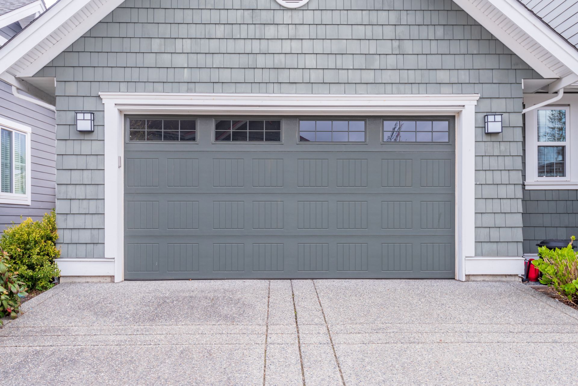 Gray garage door with windows, surrounded by gray siding, concrete driveway, and landscaping.