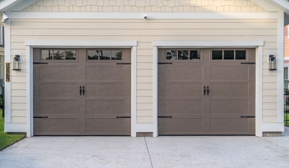 Two brown garage doors with decorative black hardware and windows on a beige house.