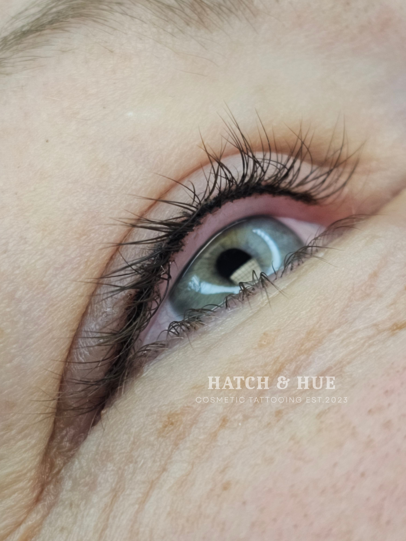 Close-up of an eye with thin black eyeliner, long lashes, and light blue iris.