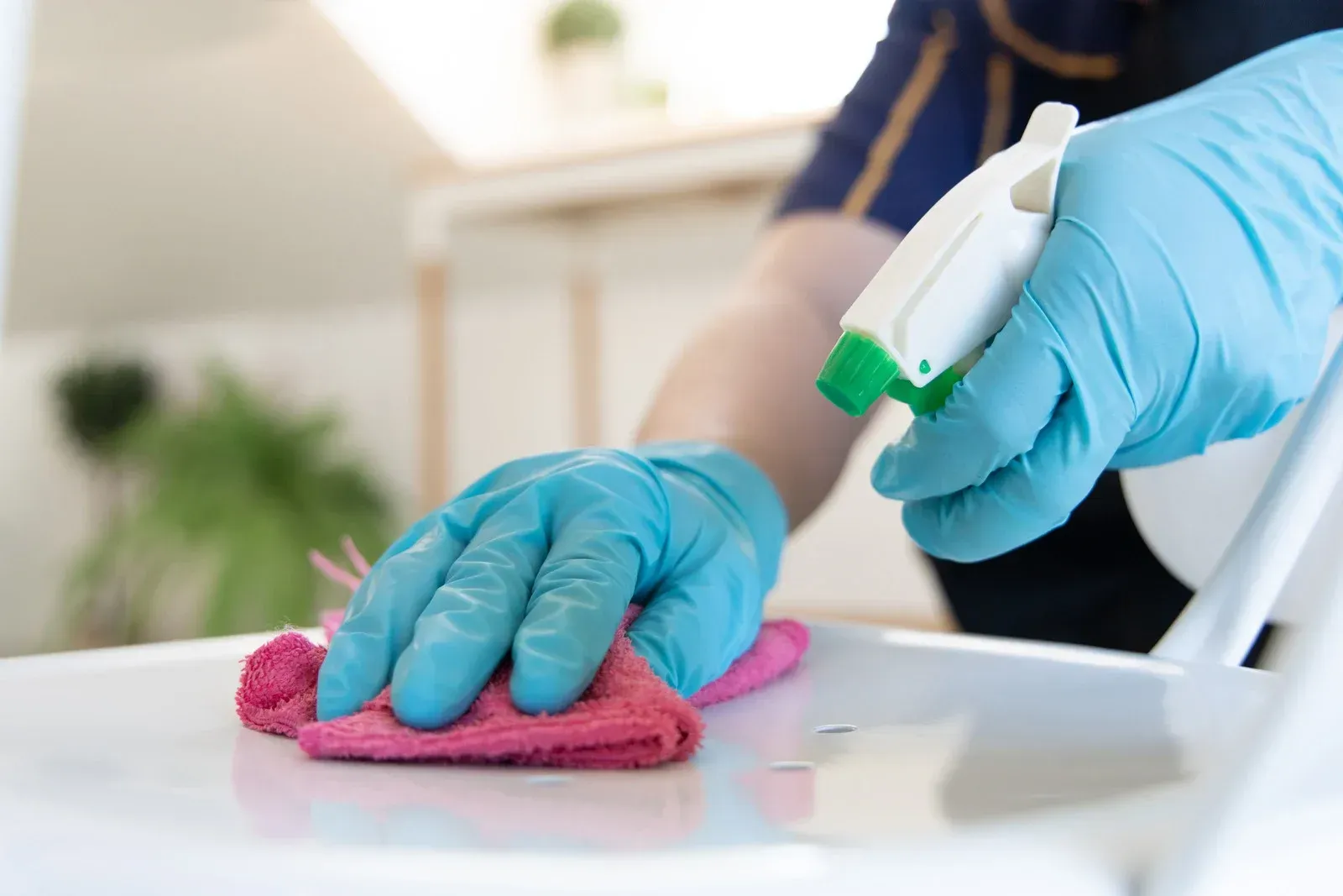 A person wearing blue gloves is cleaning a chair with a cloth and spray bottle.
