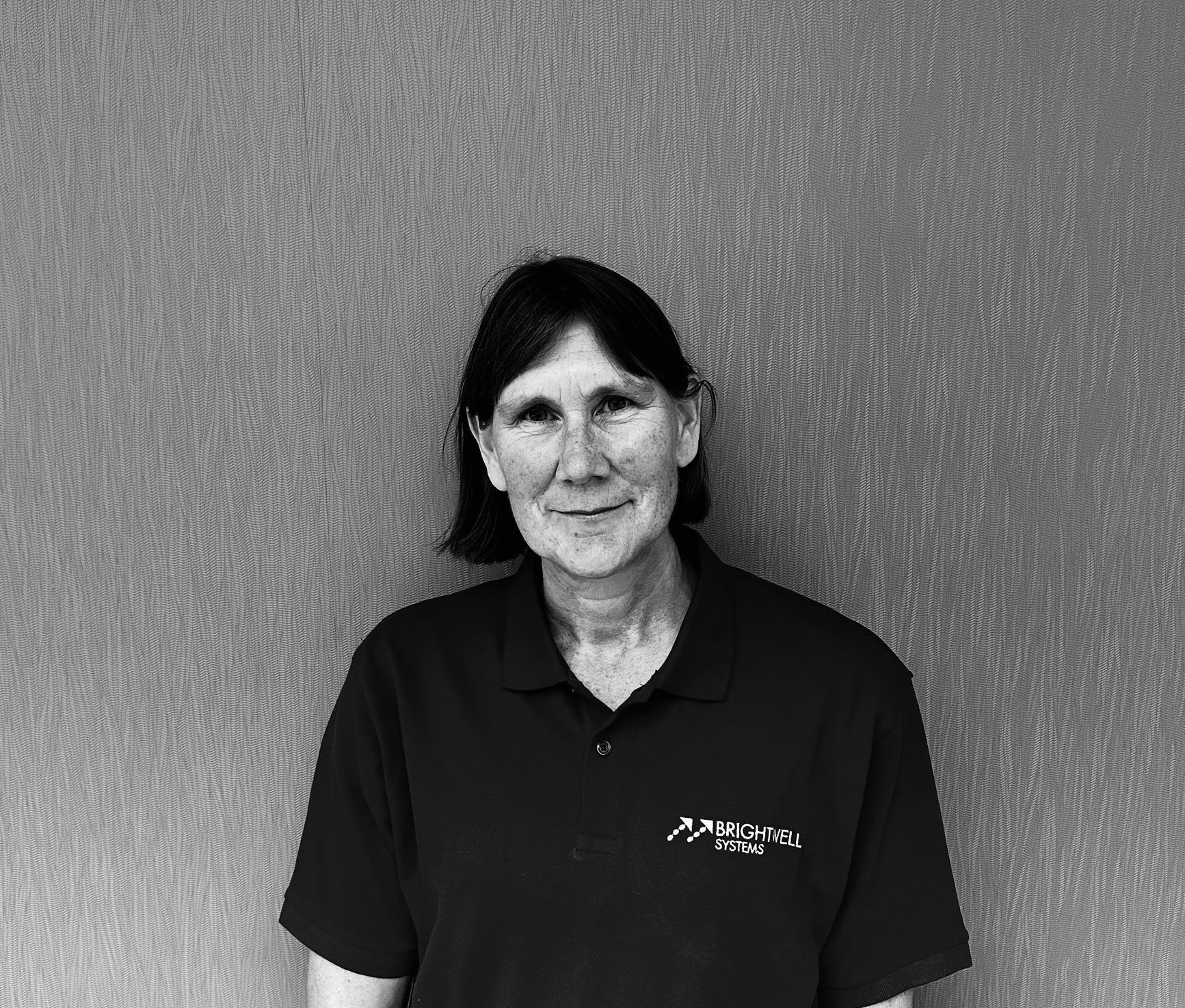 Woman in a black polo shirt smiles in front of a textured wall.