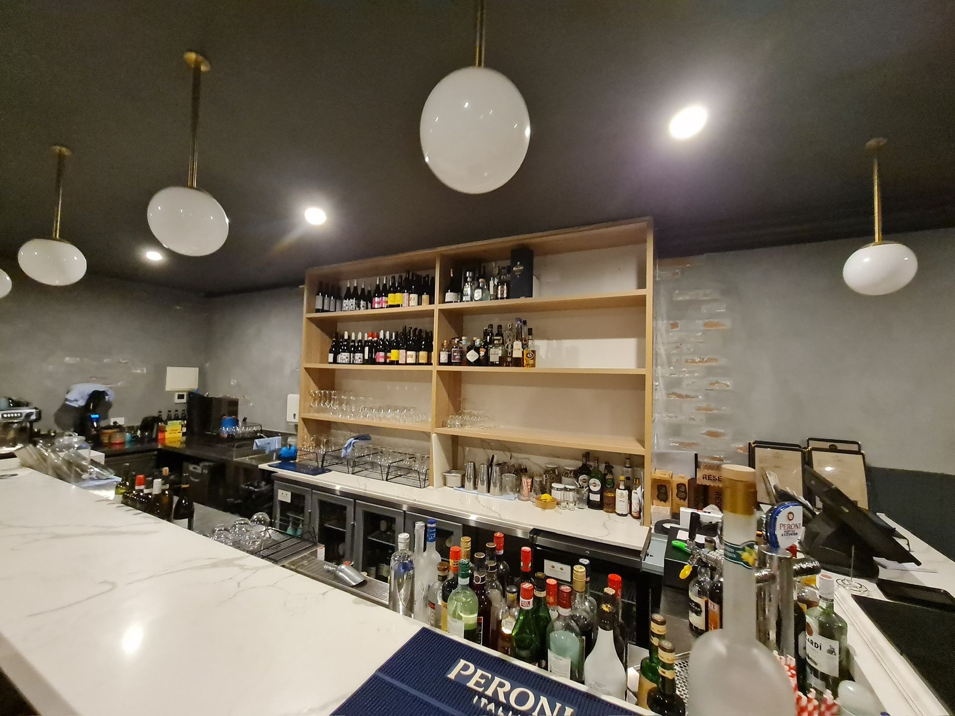Modern bar interior with white counter, liquor bottles, shelves, and hanging round lights