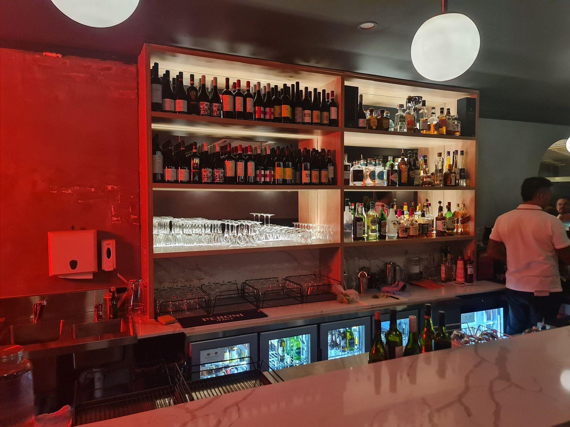 Red-lit bar with liquor shelves, glassware, and a bartender behind the counter