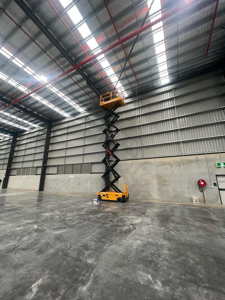 Yellow scissor lift fully extended inside a large empty warehouse with a high metal roof