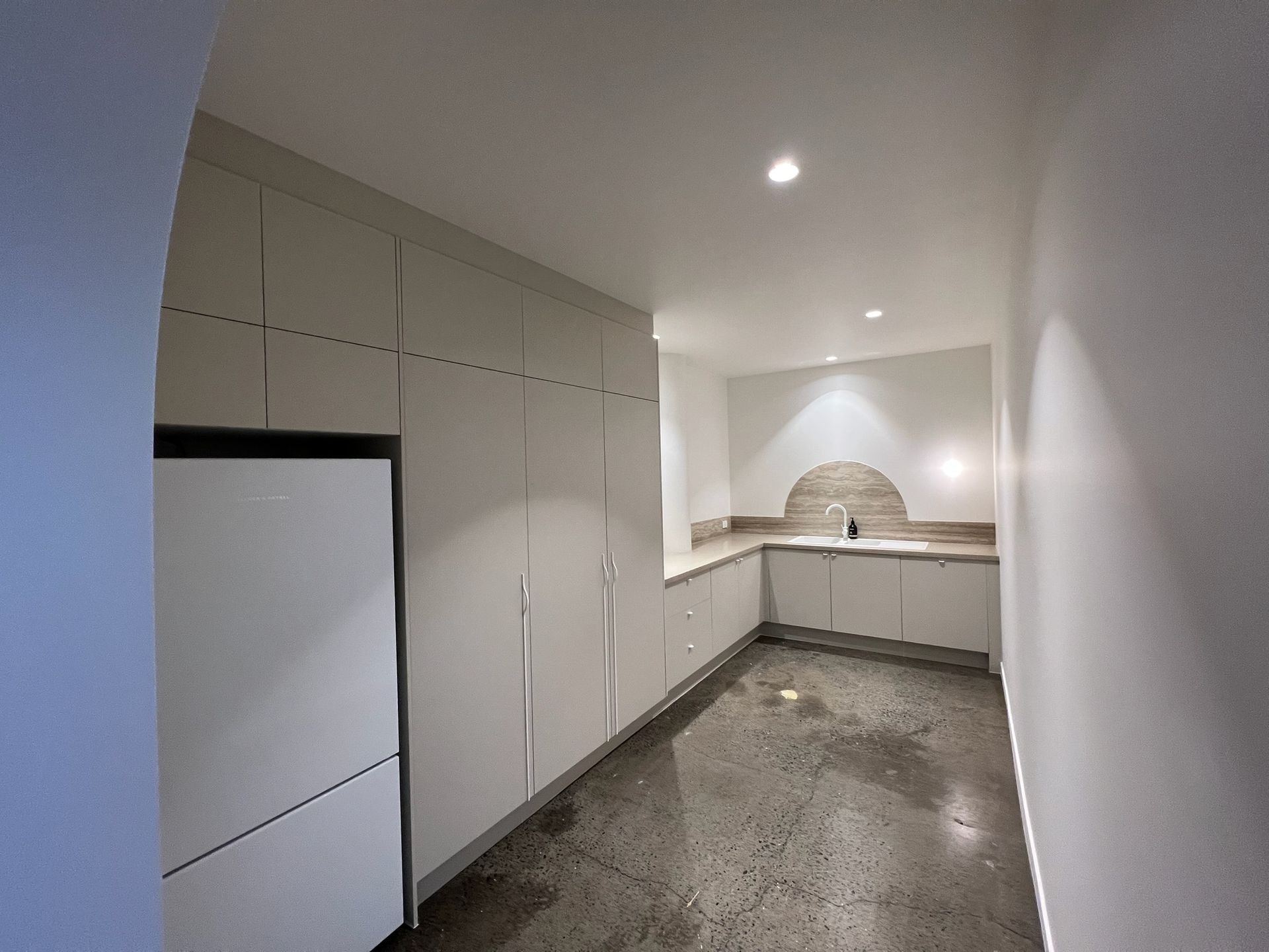Empty modern kitchen with white cabinets, built-in refrigerator, and unfinished concrete floor.