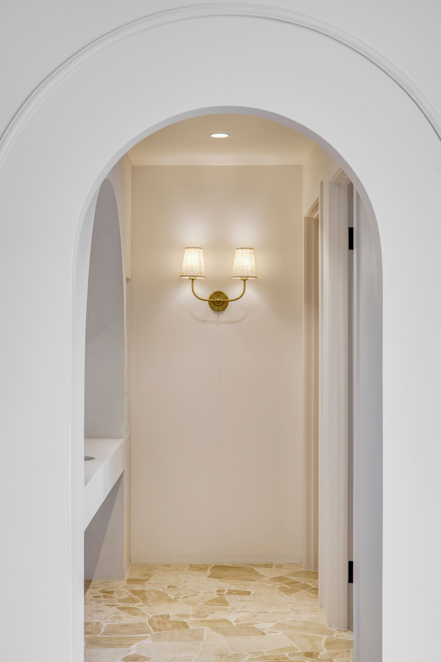 Arched hallway with cream walls, brass wall sconces, and a marble floor through a doorway