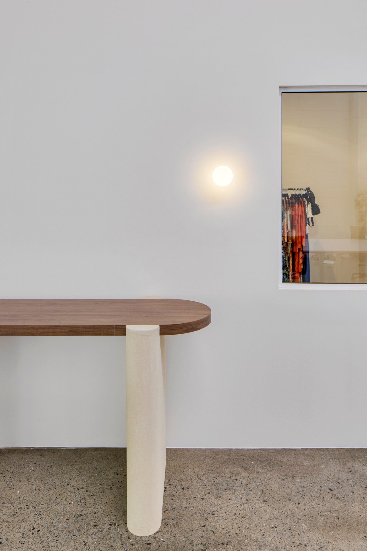 Minimalist room with a wooden table, white wall, and a small doorway showing hanging clothes.