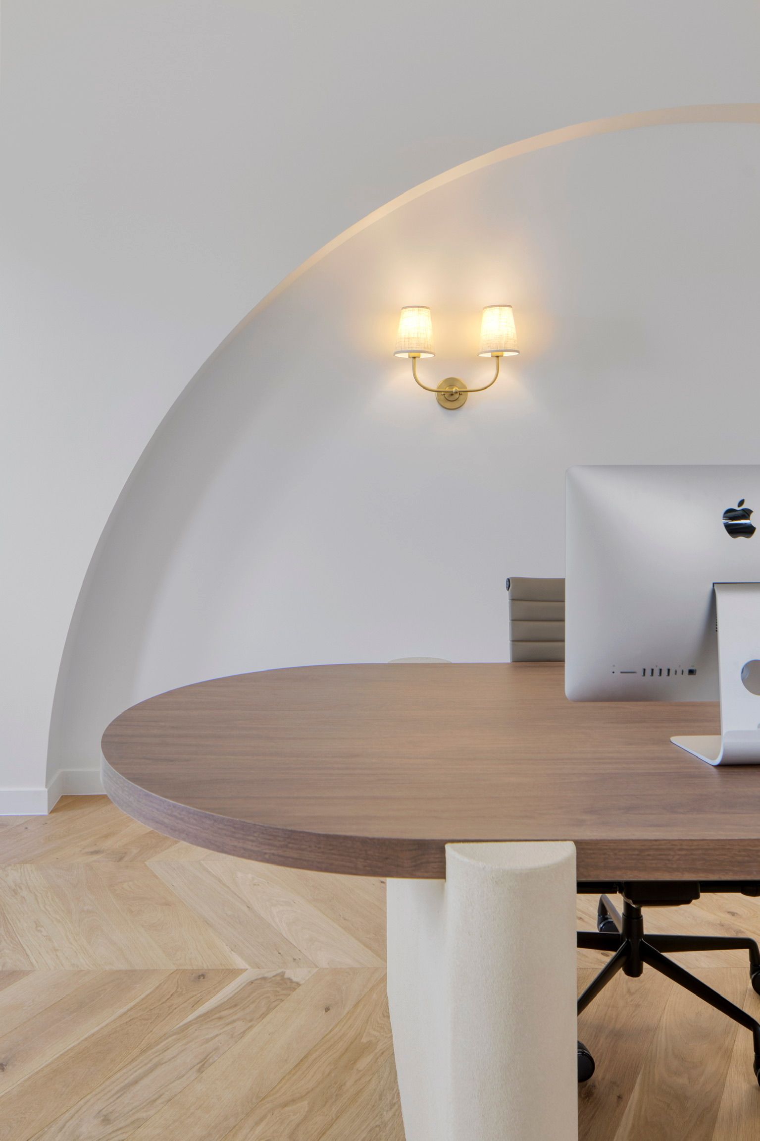 Minimalist dining area with round wooden table, white arched wall, and wall sconce lighting.