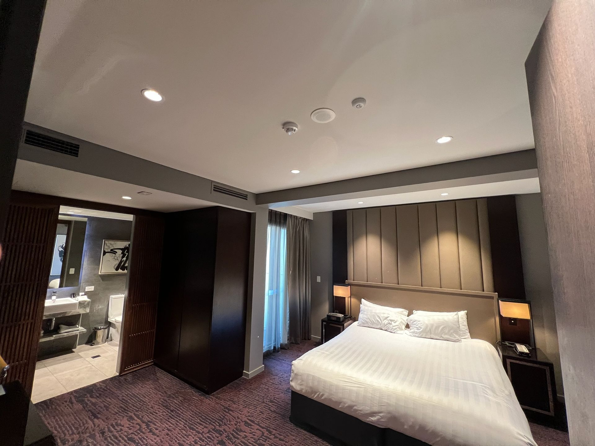 Modern hotel bedroom with white bed, dark wood accents, and a sitting area visible through the doorway