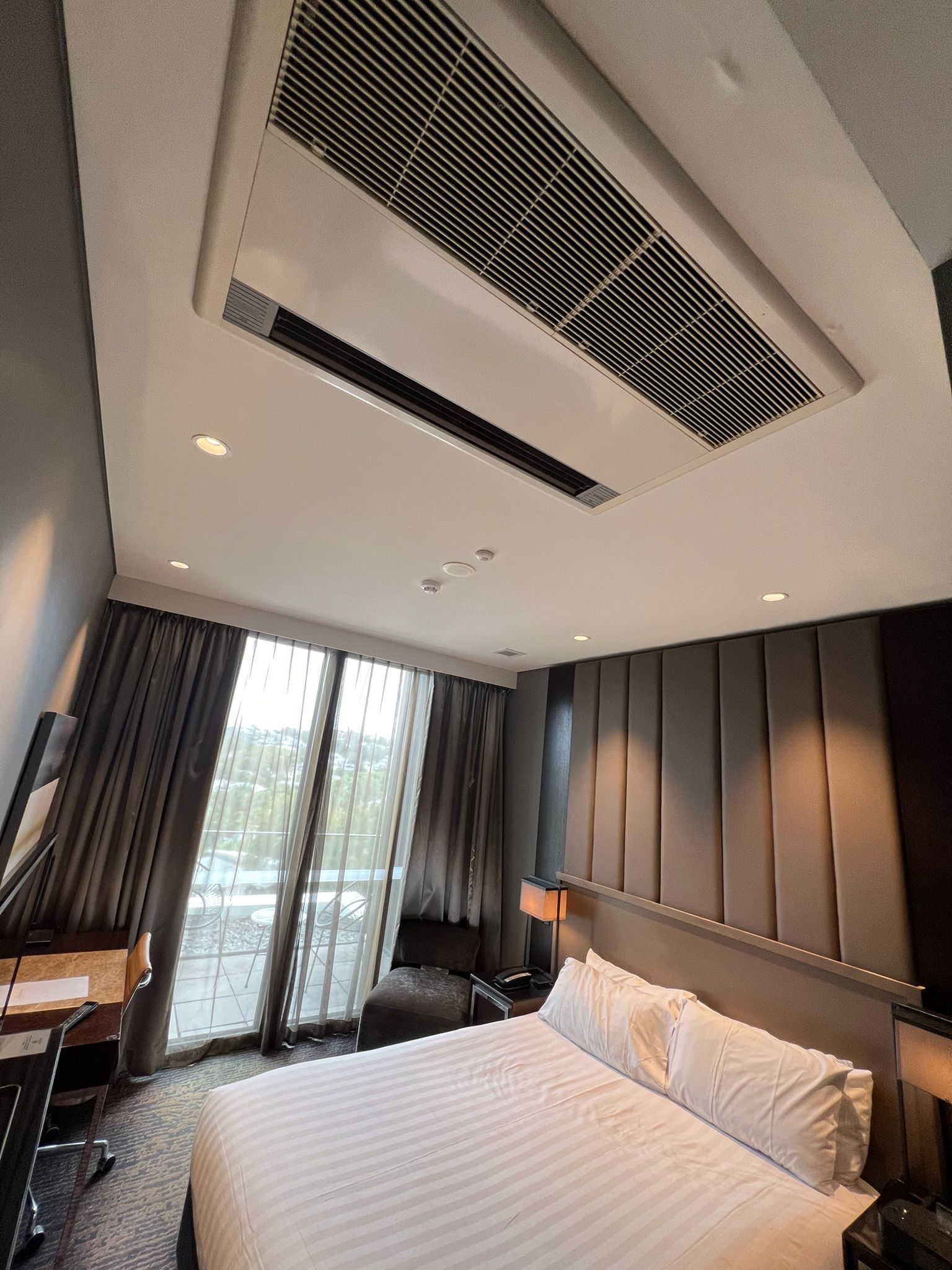 Modern hotel bedroom with a large bed, dark wood headboard, and floor-to-ceiling windows with curtains