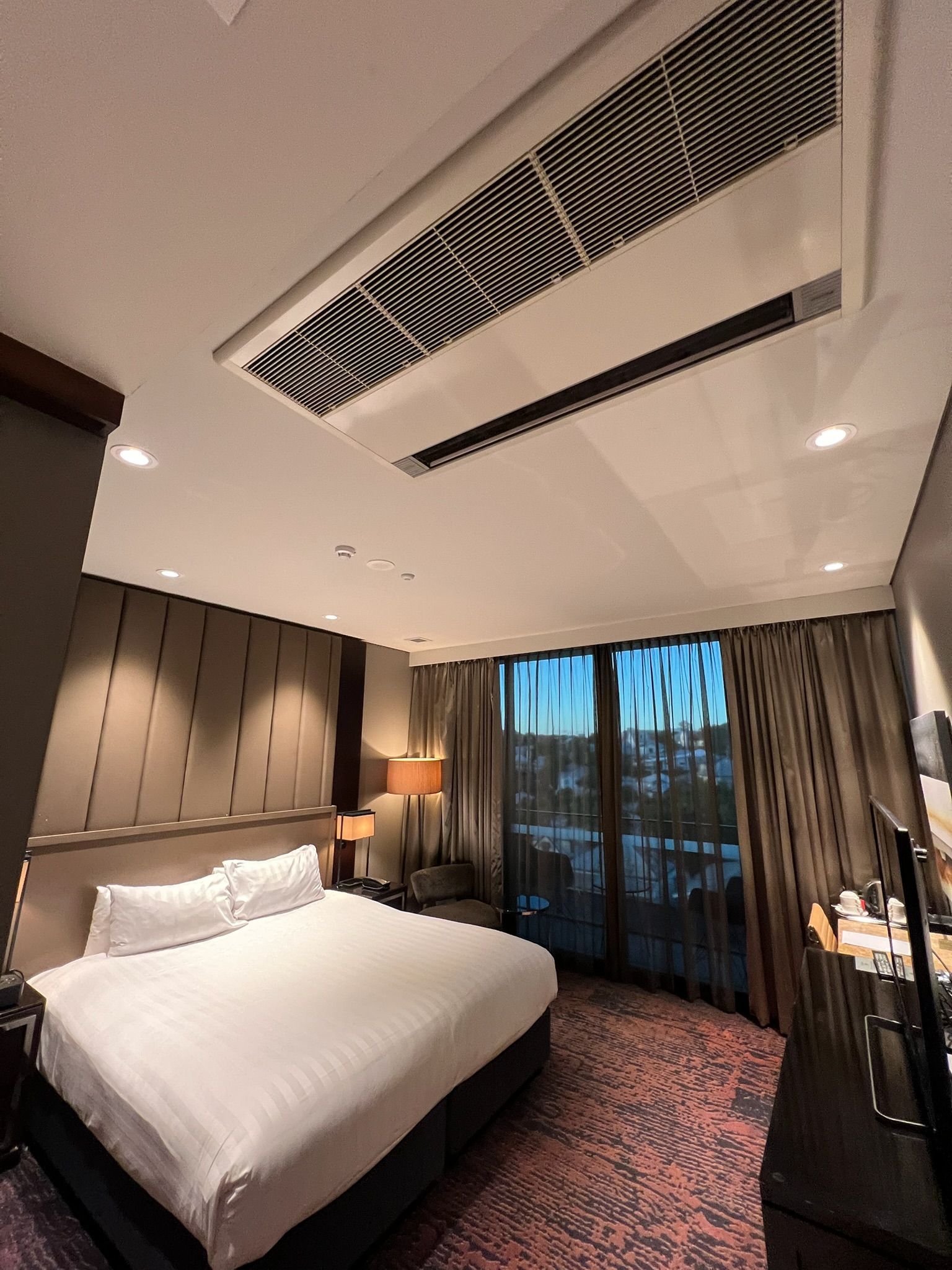 Modern hotel bedroom with large bed, dark headboard, balcony doors, and city view at dusk