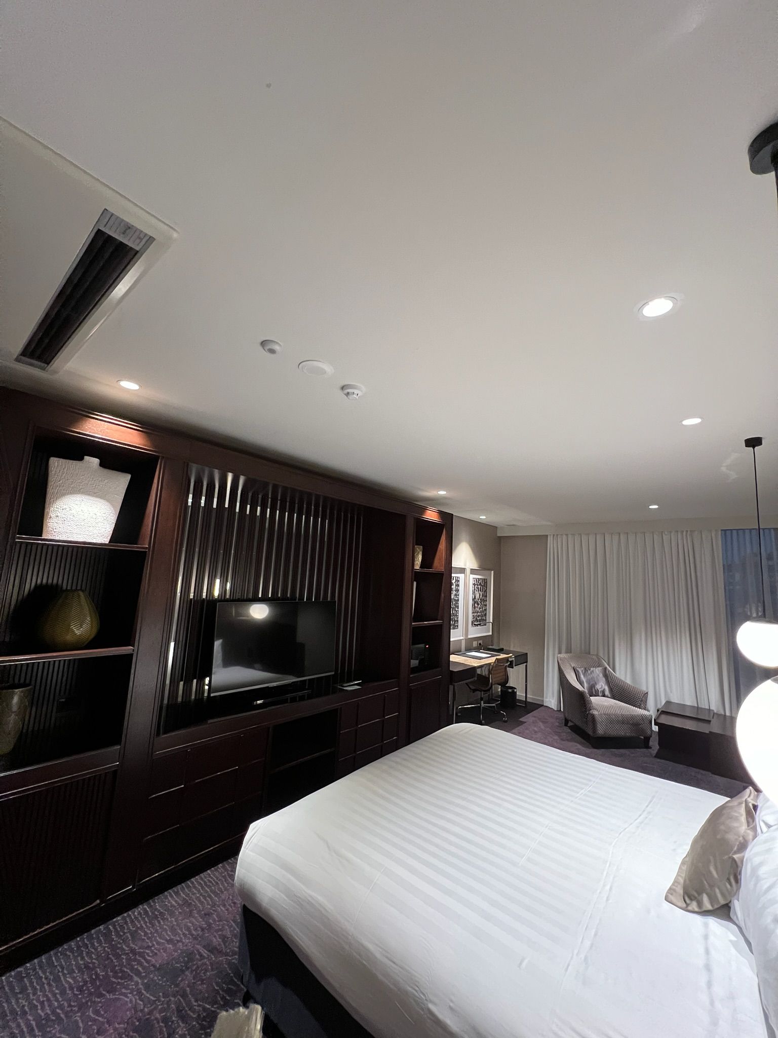 Modern hotel bedroom with a white bed, TV wall unit, curtains, and recessed ceiling lights