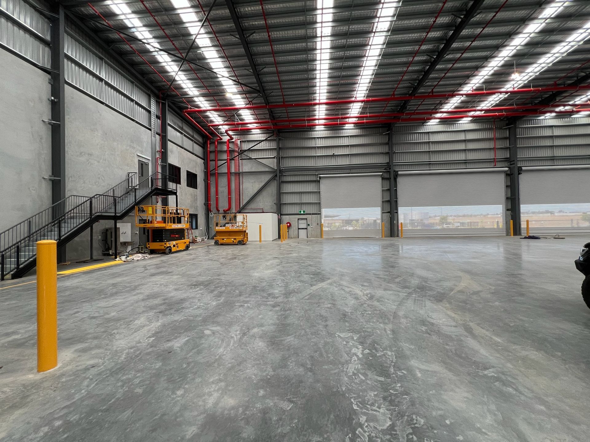 Empty industrial warehouse with concrete floor, metal walls, overhead red pipes, and yellow forklifts by the stairs