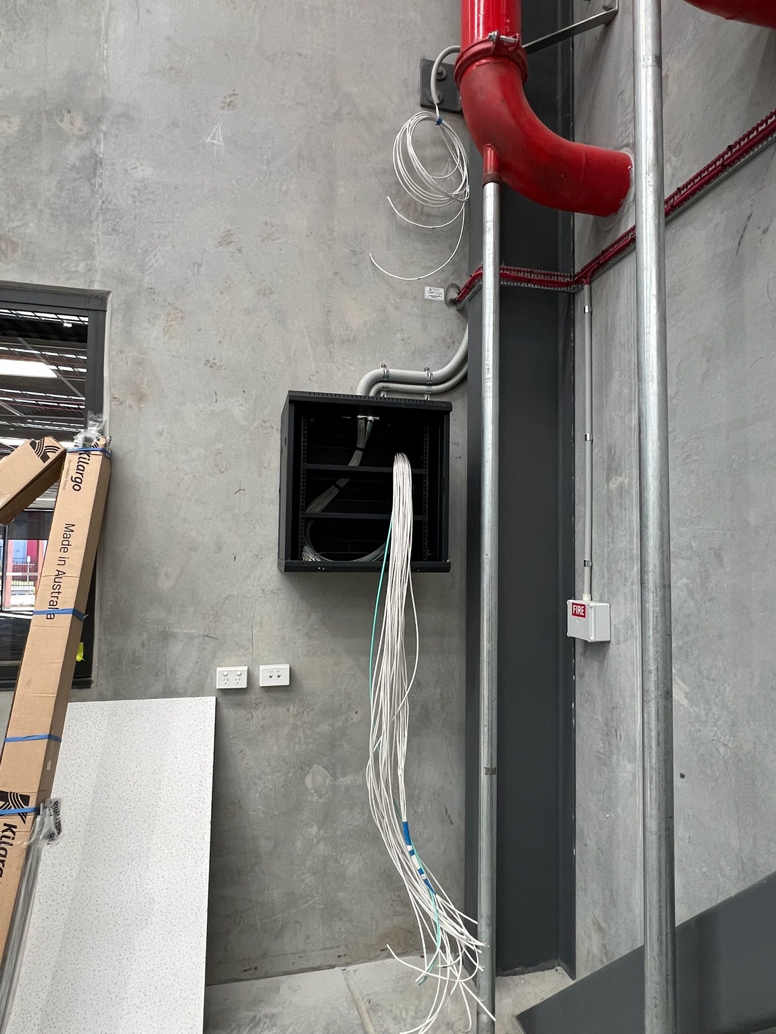 Industrial wall with black vent box and dangling white cable beneath red fire pipes.